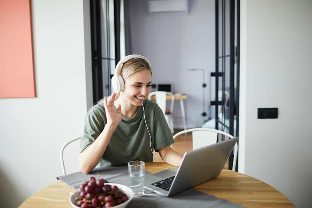 Cheerful girl in headphones waving her hand to friend while communicating through video-chat in front of laptop