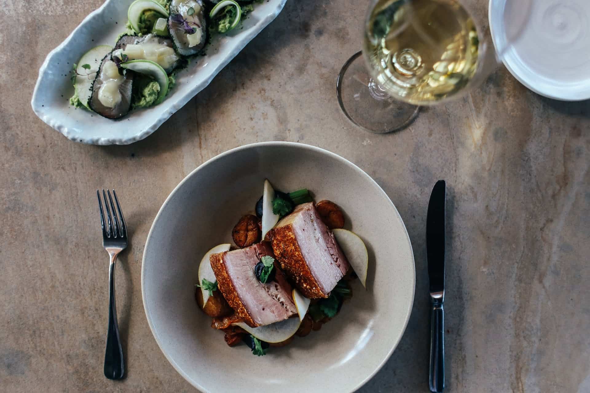 A plate of artfully placed pork in sauce with a salad and glass of wine