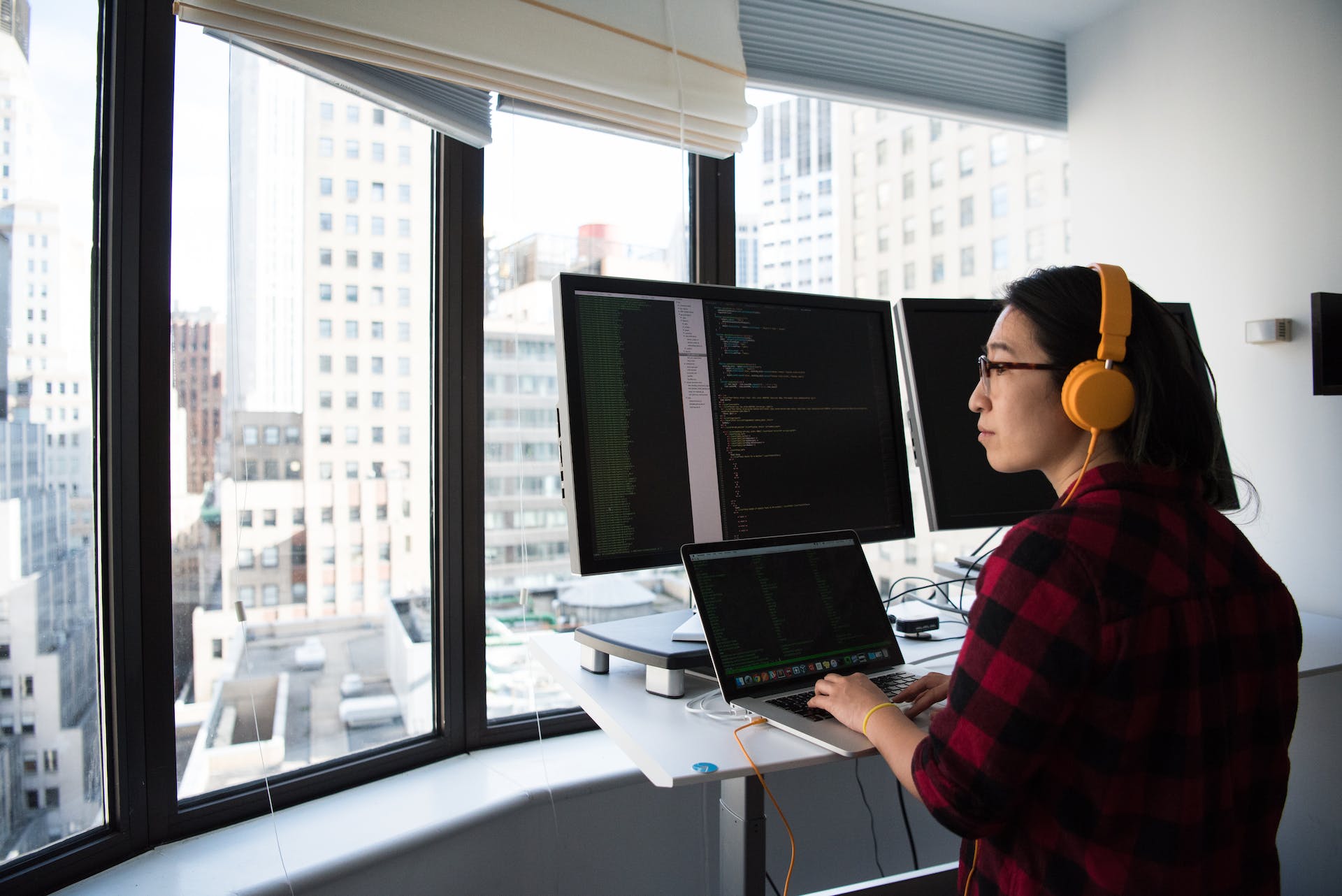 Person in red and black plaid shirt looking calmly out the window of a high rise while sitting behind three monitors displaying code