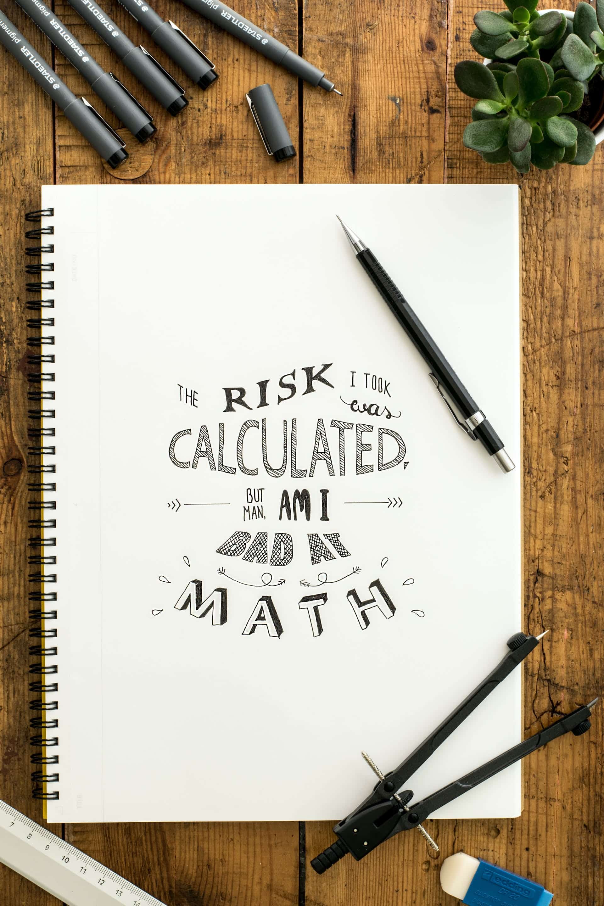 A sketch book lays on a table with pens surrounding it. Written in calligraphy on the sketch book's page is "The risk I took was calculated but man am I bad at math".