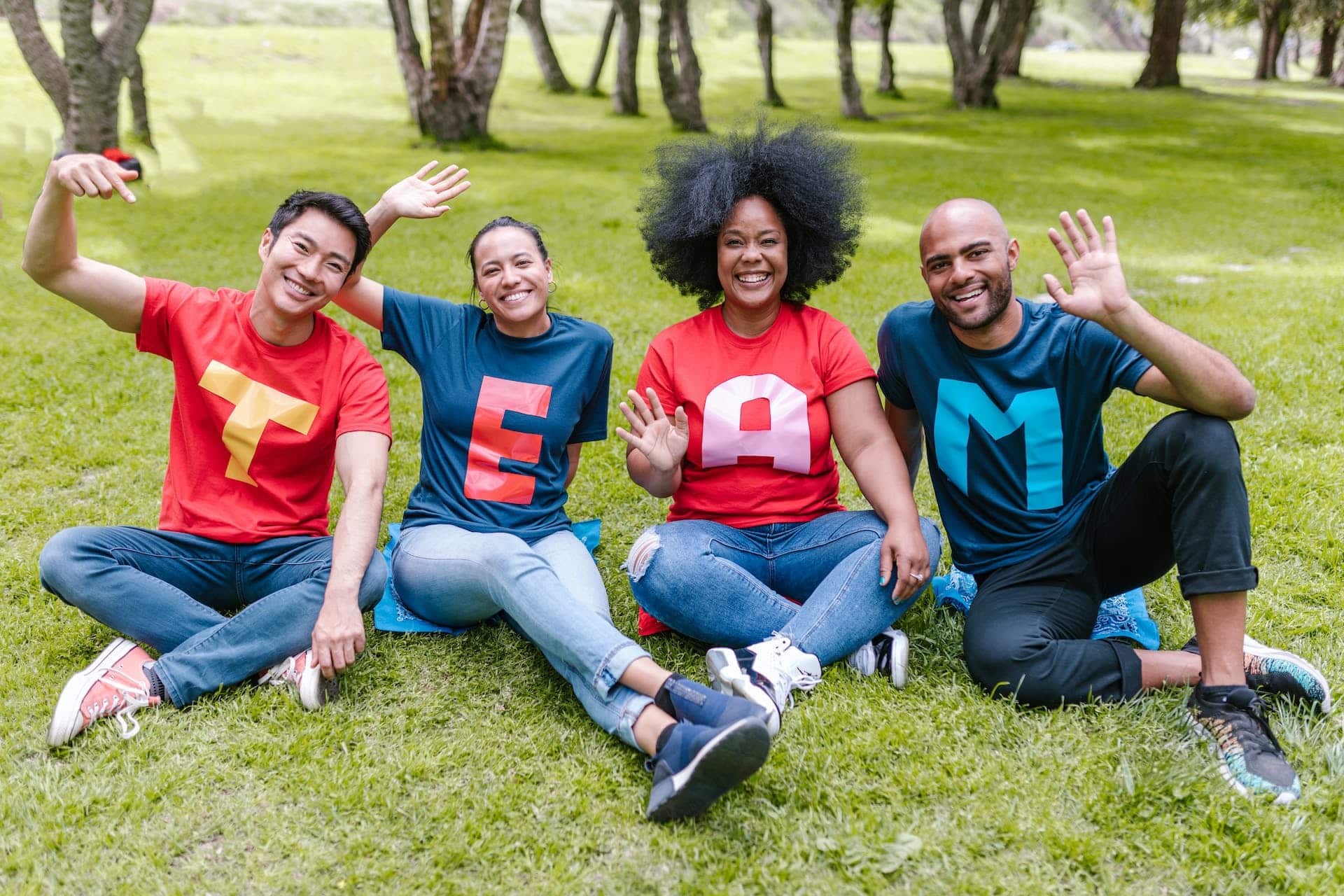 A group of four people sit in a row on a lawn while smiling and waiving. Their shirts spell out TEAM.