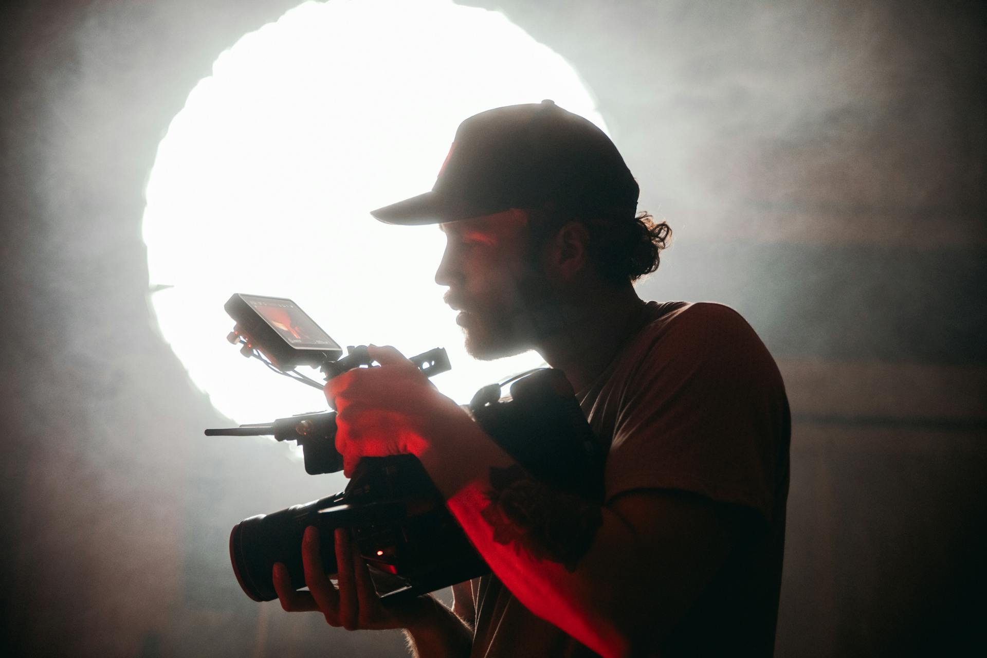 Man wearing a cap is focused on a camera while filming. He is front lit in red with a bright white backlight.