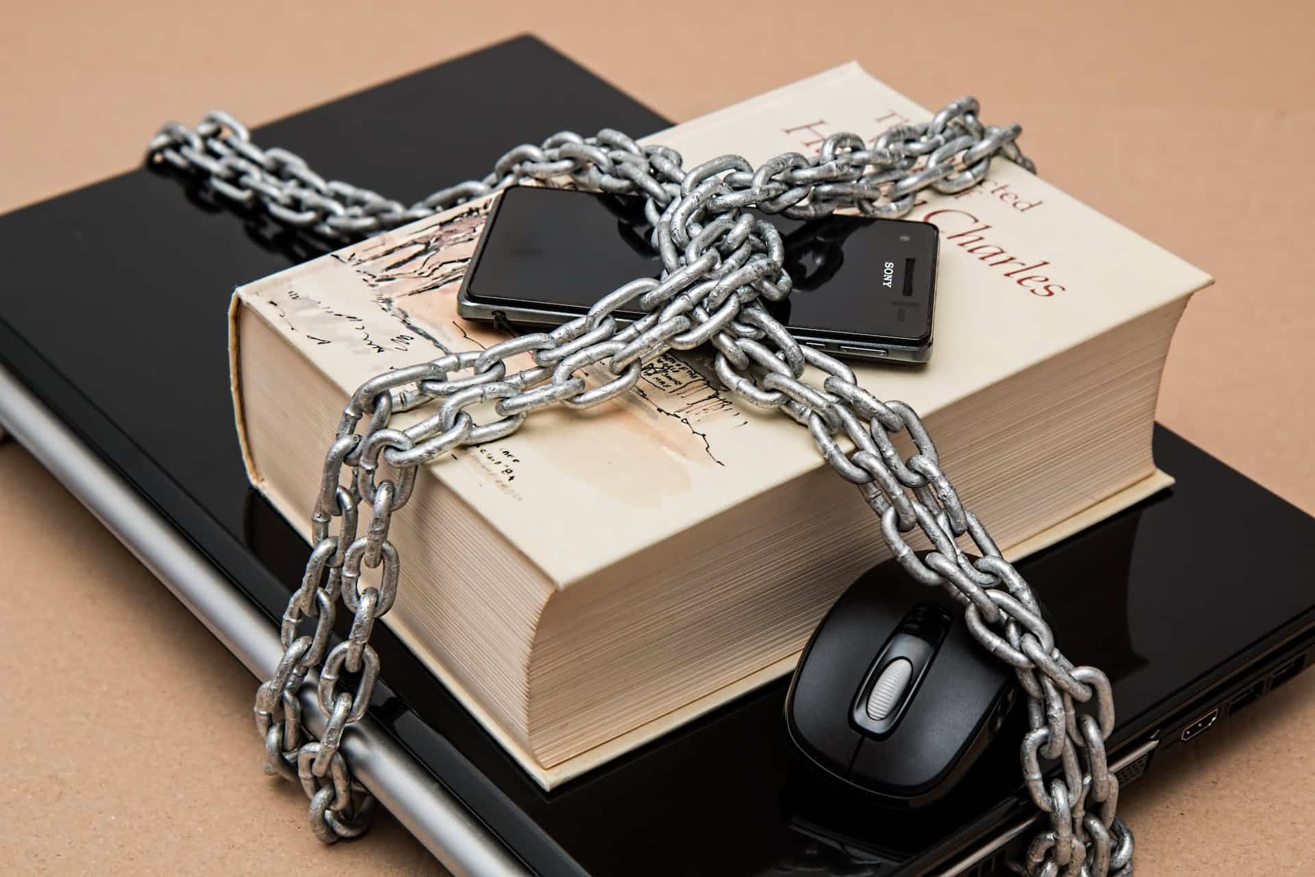 A laptop, book, phone, and computer mouse chained together with a thick chain.