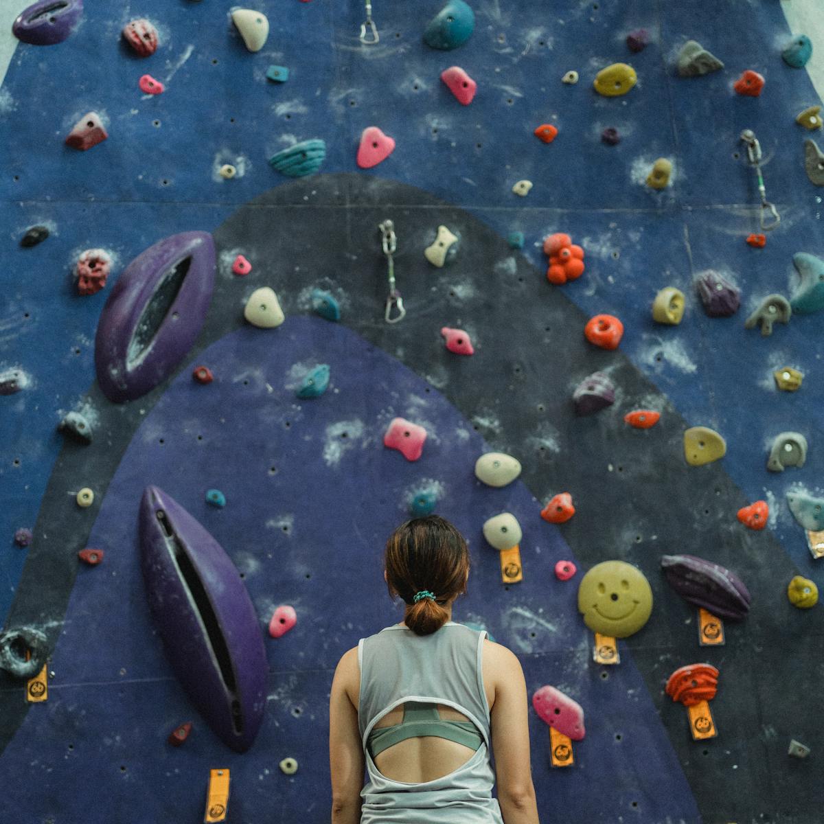 Woman in gym clothes stands with her back to the camera staring up at the base of a colorful climbing wall.