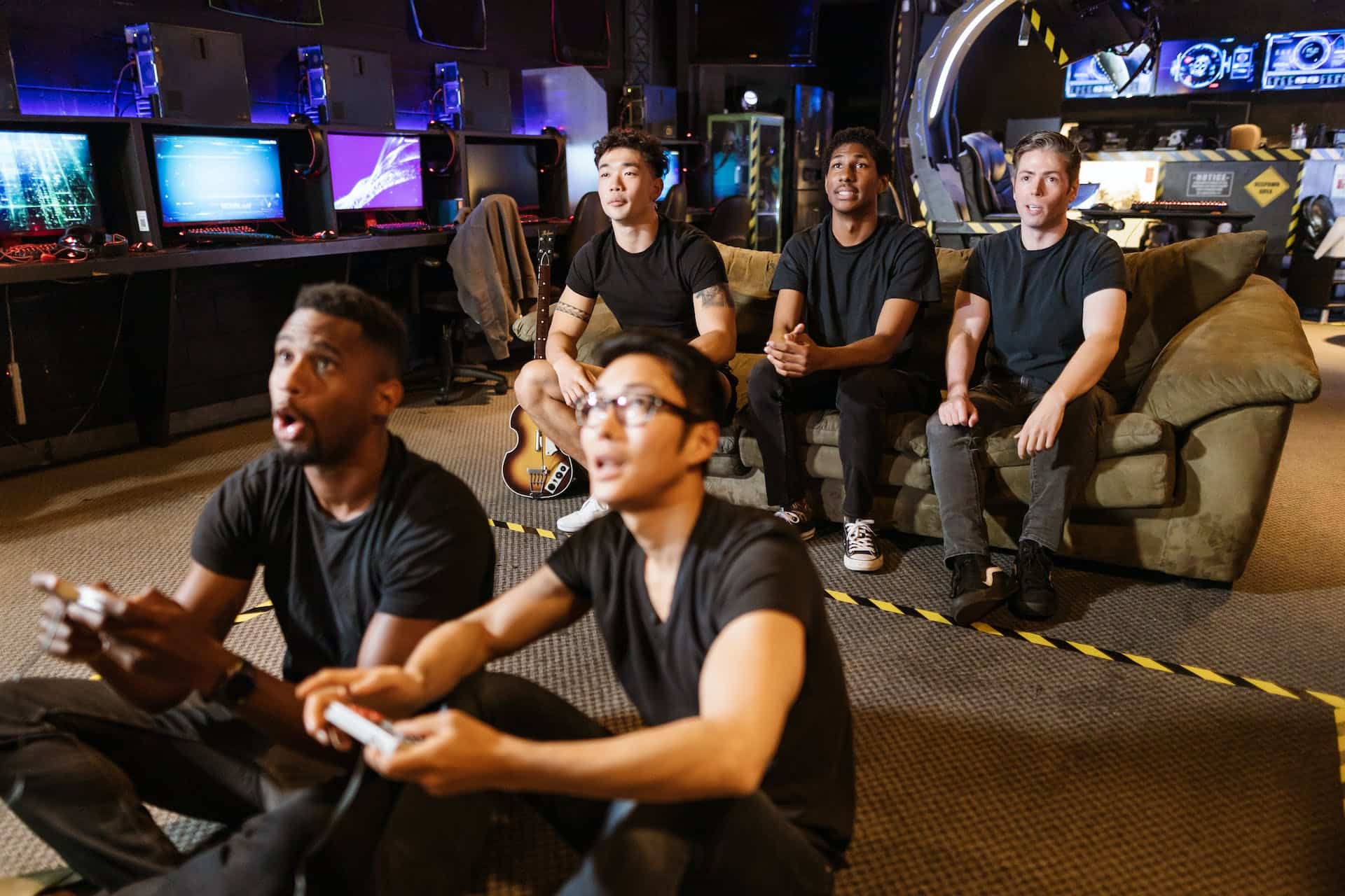 Two young men are engrossed in playing a video game while three young men raptly watch from a couch behind them.