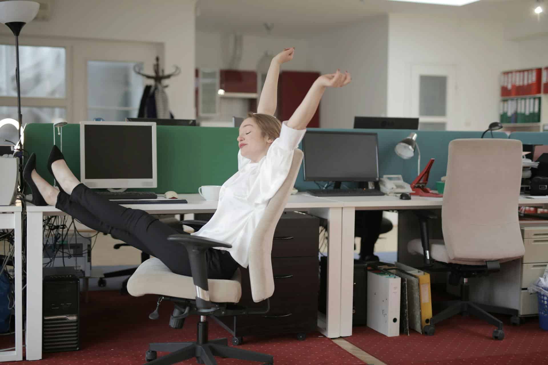 Woman stretches arms widely while sitting at an office desk with feet kicked up on desk.