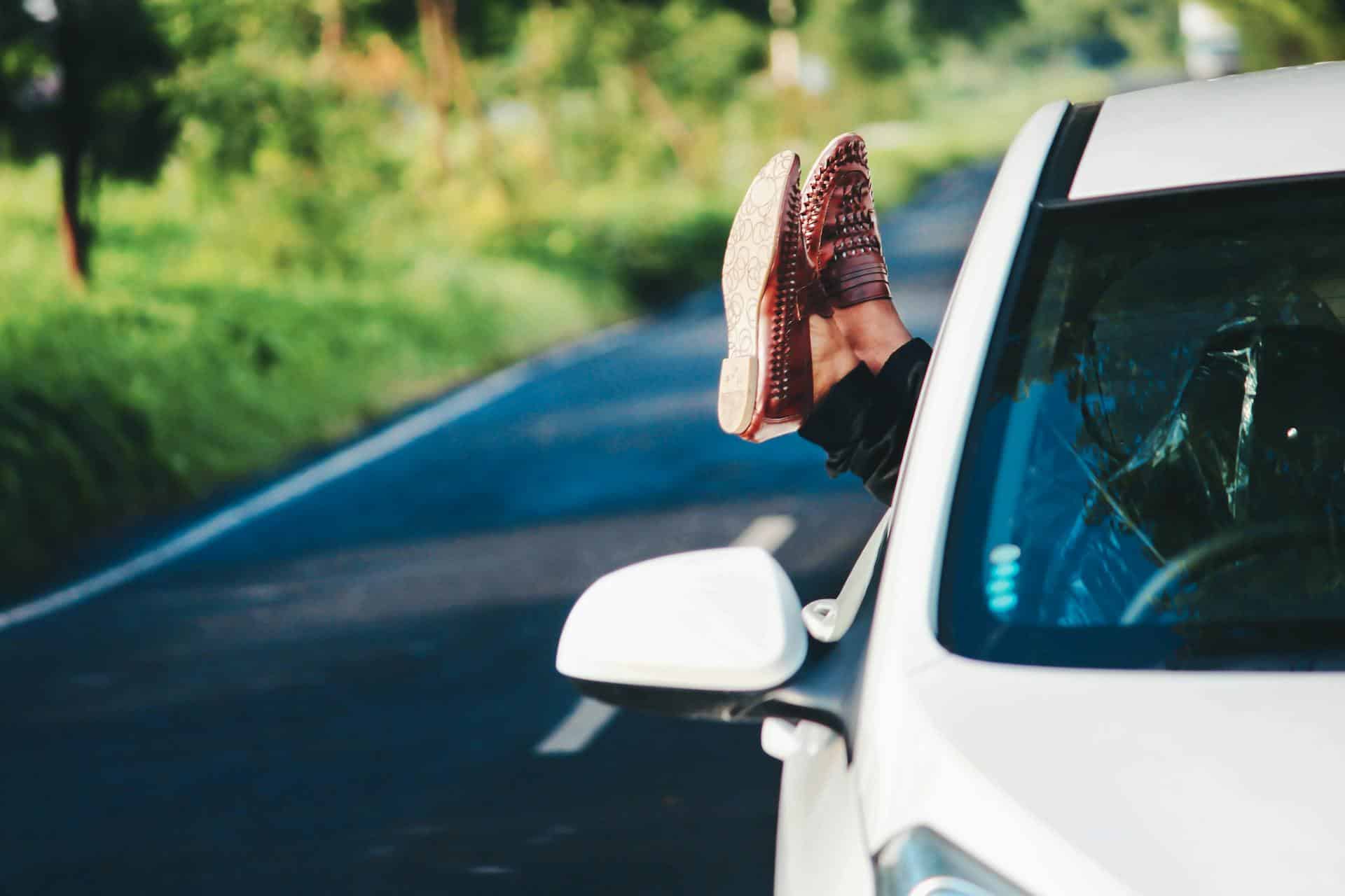 A pair of feet hang out the driver's side of a car while it drives down a road.