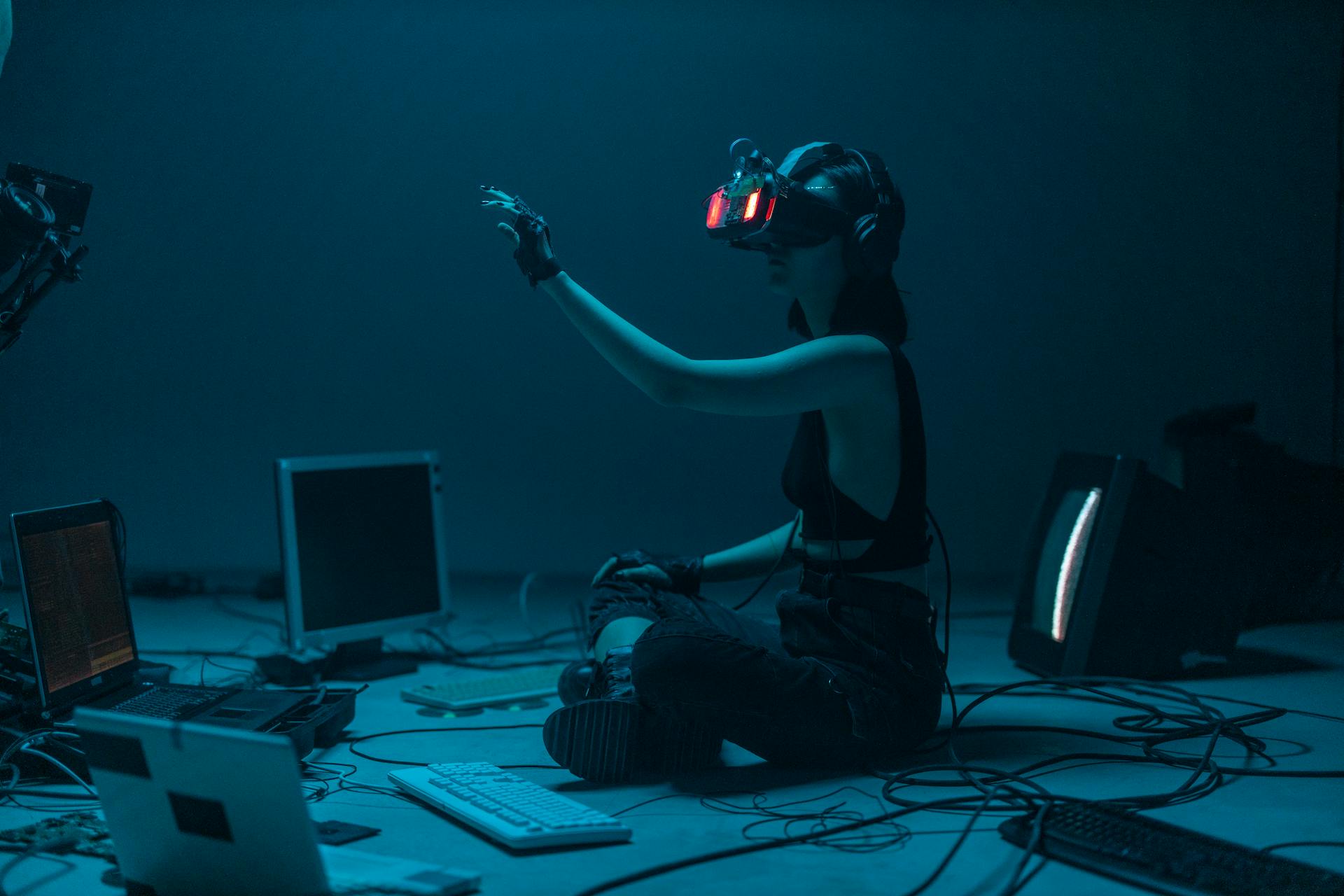 Kneeling woman surrounded by monitors wears glowing red VR goggles while reaching out to something the viewer cannot see