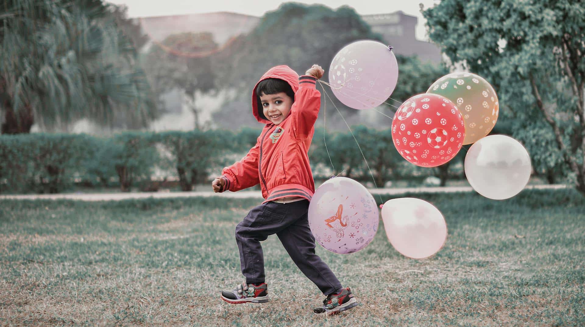 Child in a red sweatshirt grins at the camera while running through a park with large colorful balloons trailing behind him.