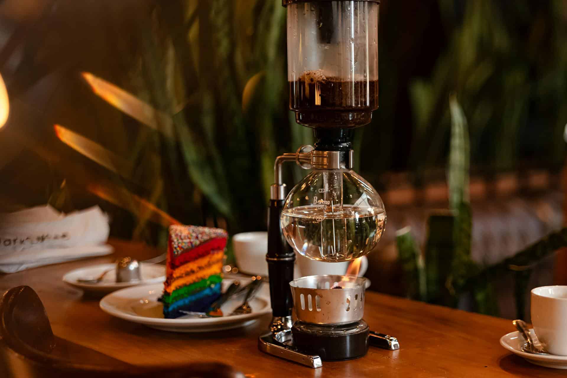 Image of a coffee syphon sitting on a table with a piece of rainbow cake in the background.