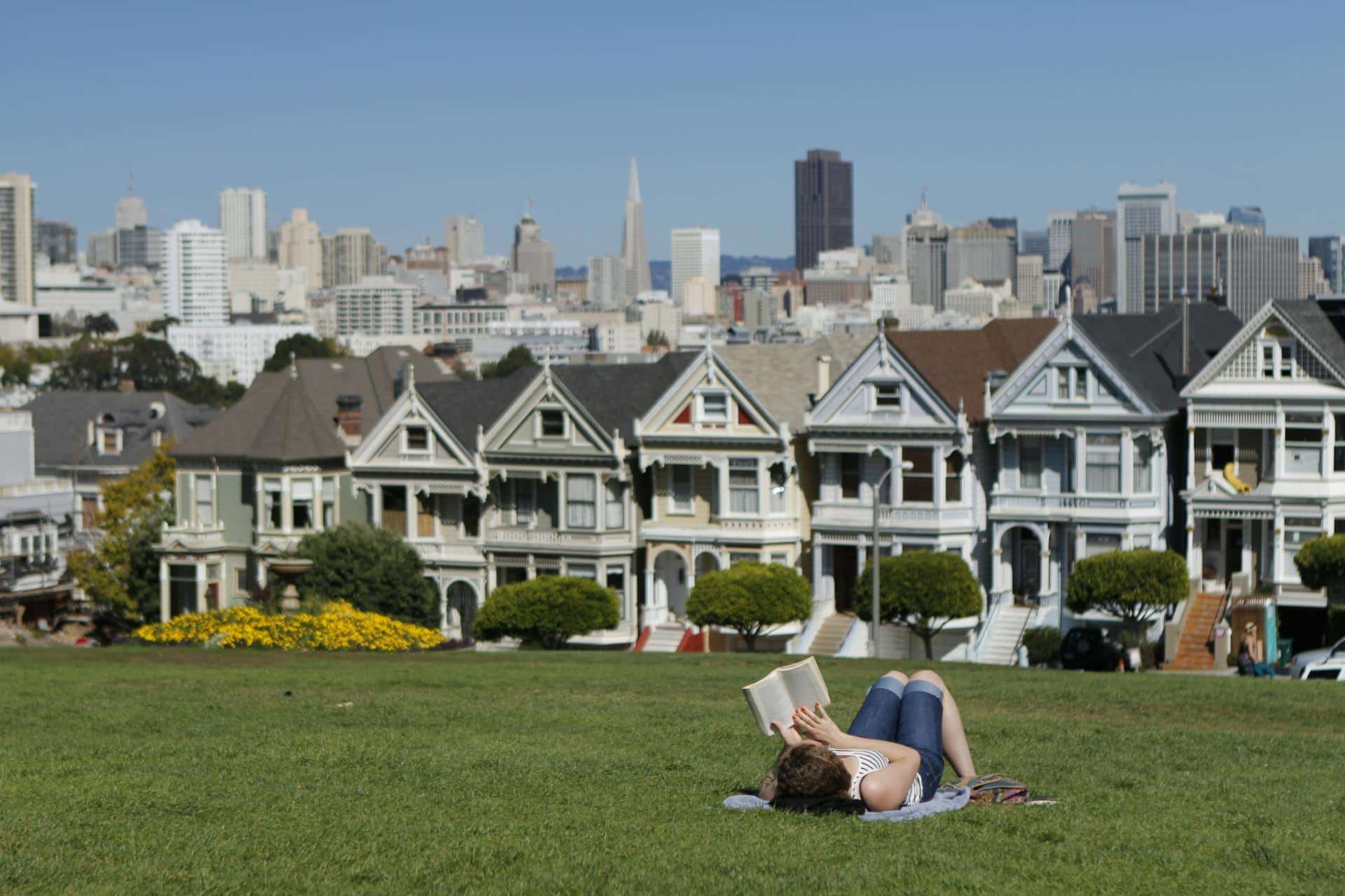 Woman lies down on a picnic blanket reading a book under a bright sun in Alamo Square Park.