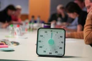 A clock sits in the foreground while a work group is heads down in the background.