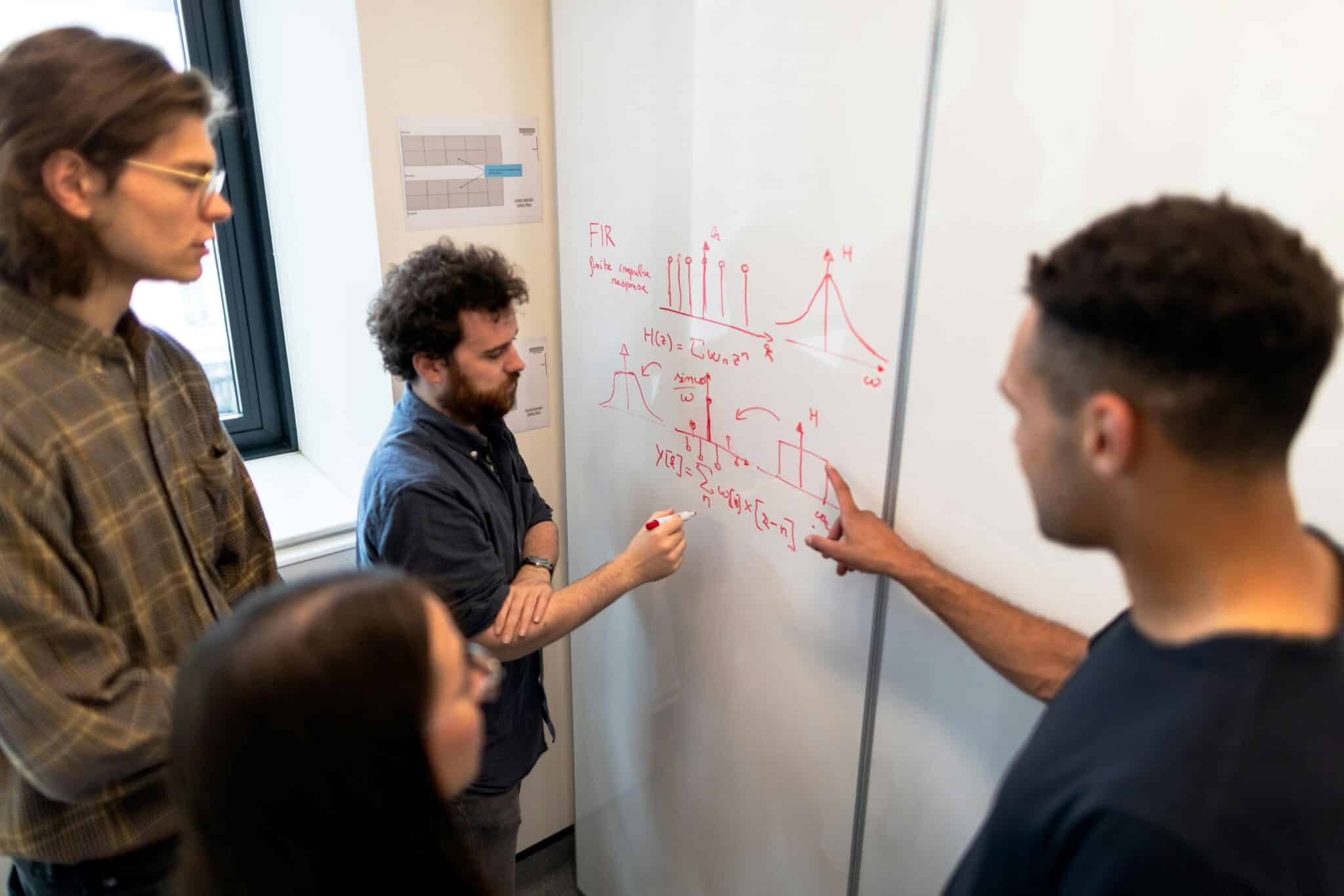 People gathered around a whiteboard discussing equations and graphs drawn on the whiteboard.