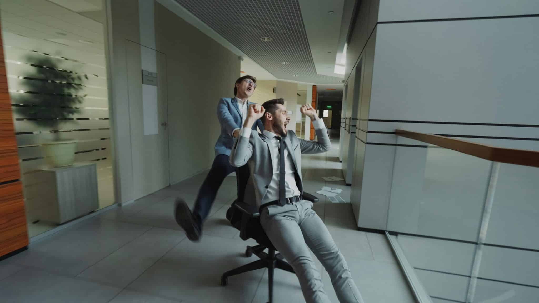 A man pushing an office chair down the hall with another man sitting in it